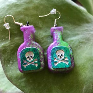 May include: A pair of earrings shaped like poison bottles. The earrings are purple and green with a skull and crossbones design on a green background. The earrings have gold-colored hooks.