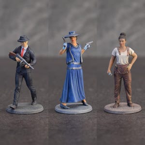 May include: Three miniature figurines standing on circular bases. The first is a person in a dark suit, fedora, and red tie holding a submachine gun. The second is a person in a blue dress and hat holding two pistols. The third is a person in overalls.