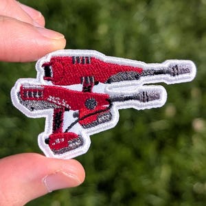May include: An embroidered patch featuring a red and grey power drill design with a white border. The patch depicts a cordless drill with a battery pack at the base and a drill bit extending forward, rendered in detailed red and grey thread work.