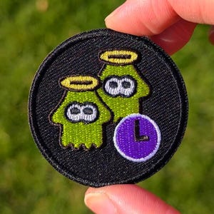 May include: A round, black embroidered patch featuring two green cartoon characters with yellow halos and the number eight. A purple circle with a white "L" is on the right. The patch is held by a hand against a blurred green background.
