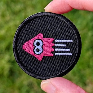 May include: A circular, black embroidered patch with a pink squid design, the number 8, and three white lines. The patch is held between fingers against a blurred green background.