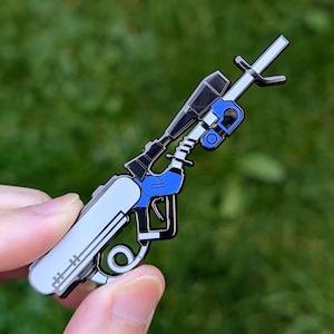 May include: A detailed enamel pin depicting a futuristic rifle with a scope. The pin features a color scheme of white, black, and blue. The design includes intricate details, such as the scope, trigger, and other mechanical elements. The pin is held against a green background.