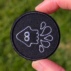 May include: A round, black embroidered patch featuring a white outline of a stylized squid with the number 8 and water droplets. The patch is held up against a green, grassy background.