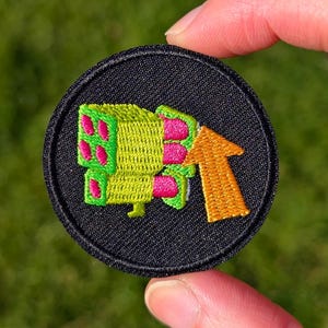 May include: Embroidered patch with a black circular base. The patch features a neon green and pink design of a stylized weapon with an orange arrow. The patch is held by a hand against a blurred green background.
