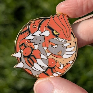 Puede incluir: Un pin redondo de esmalte dorado con una criatura roja y naranja con detalles blancos y un ojo amarillo. El pin se sostiene entre dos dedos sobre un fondo verde borroso.