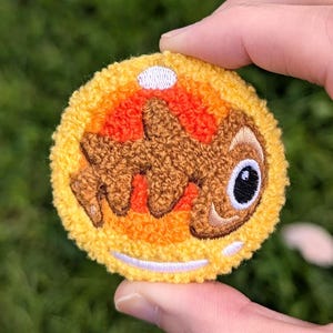 Könnte beinhalten: Gestickter Aufnäher mit einem Cartoon-Charakter mit braunem Körper, schwarzem Auge und gelb-orangem Hintergrund. Der Aufnäher hat eine flauschige Textur und einen weißen Rand. Der Aufnäher wird von einer Hand vor grünem Hintergrund gehalten.