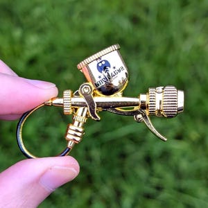 May include: A miniature gold spray gun with a black hose and a small cup. The cup features a cartoon character and the text "King Lize". The spray gun is set against a green grassy background.