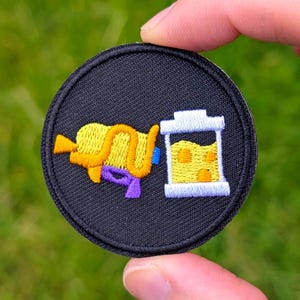May include: Embroidered patch with a black background featuring a yellow and purple water gun and a white and yellow container. The patch is round and held by a hand against a green background.