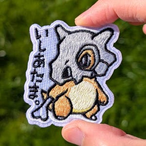 May include: Embroidered patch featuring a cartoon Cubone character with a bone helmet and a tan body. The patch has Japanese text and a white border. The patch is held against a green background.