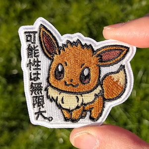 May include: An embroidered patch featuring a brown and cream-colored Eevee character with large eyes and pointed ears. To the left of the character, the Japanese text "可能性は無限大。" is embroidered in black thread on a white background.