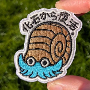May include: An embroidered patch featuring a brown ammonite fossil with a spiral shell and bright blue tentacles. The patch has a white border and includes the Japanese text 化石から復活 written in black thread along the top edge of the design.
