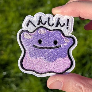 May include: An embroidered patch featuring a purple, amorphous, blob-like character with two small black eyes and a simple black line for a mouth. Above the character, the Japanese text "へんしん!" is stitched in black thread on a white background.