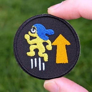 May include: Round black embroidered patch featuring a yellow figure with blue hair running towards a yellow arrow pointing upwards. The patch also includes three white lines below the figure. The patch is held in a hand against a green background.