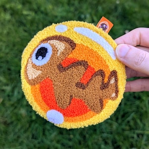 Pode incluir: Um patch bordado redondo com um desenho de peixe de desenho animado. O patch é predominantemente amarelo, laranja e marrom, com um acabamento texturizado. O peixe tem um olho grande e um corpo detalhado. O patch é segurado contra um fundo de grama verde.