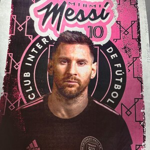 May include: A rectangular poster featuring a portrait of Lionel Messi in a black jersey. The background is vibrant pink with black geometric patterns and the text Messi 10, Club Internacional de Fútbol Miami, and the Adidas logo are clearly visible.