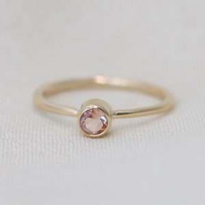 May include: A delicate gold ring with a single, round, light pink gemstone.