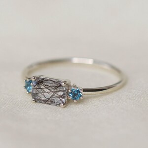 May include: A silver ring featuring a rectangular, clear gemstone with black inclusions, flanked by two small, round, blue gemstones. The ring is set against a soft, off-white background.