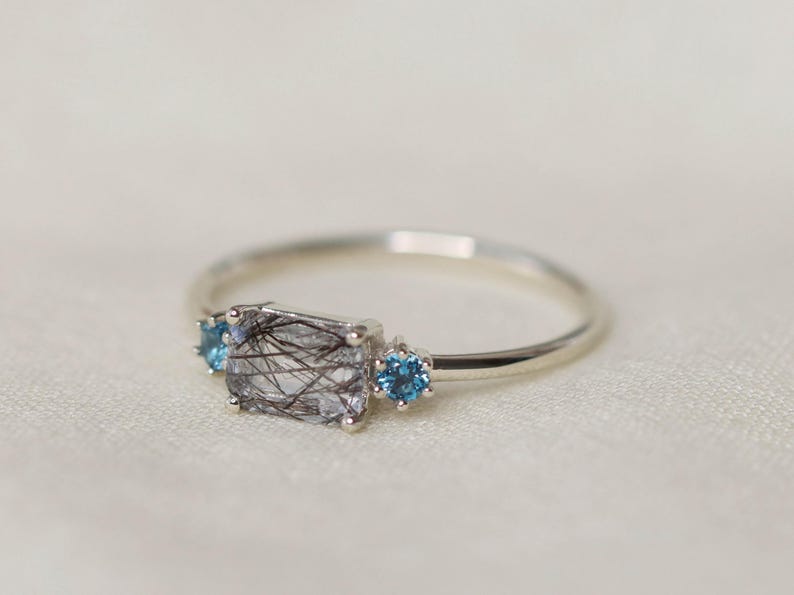 May include: A silver ring featuring a rectangular, clear gemstone with black inclusions, flanked by two small, round, blue gemstones. The ring is set against a soft, off-white background.