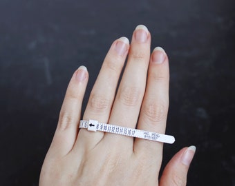 Könnte beinhalten: Ein weißer Kunststoff-Ringgrößenmesser mit den Zahlen 7 bis 17 darauf gedruckt. Der Größenmesser befindet sich an einem Finger. Der Text "PAT. PEND. 9101687" ist auf dem Größenmesser aufgedruckt.