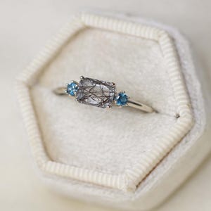 May include: A silver ring featuring a rectangular, clear gemstone with black inclusions, flanked by two small, round, blue gemstones. The ring is set on a white velvet surface, showcasing the jewelry's design and details.