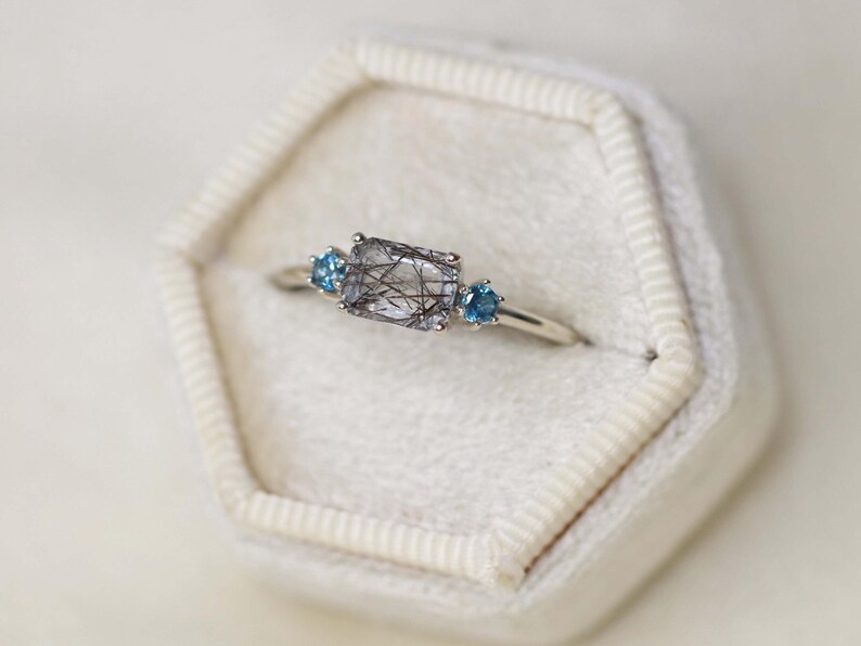 May include: A silver ring featuring a rectangular, clear gemstone with black inclusions, flanked by two small, round, blue gemstones. The ring is set on a white velvet surface, showcasing the jewelry's design and details.