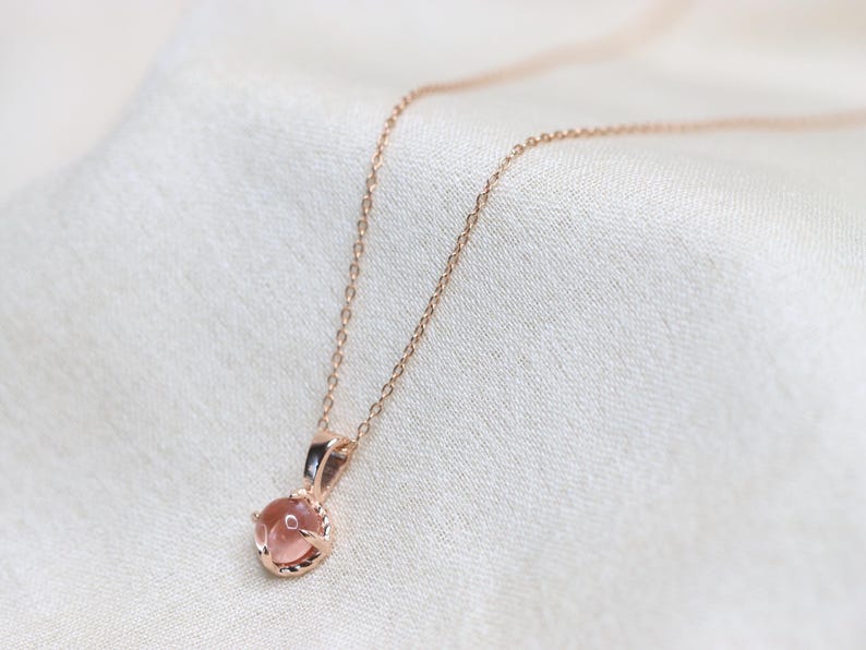 May include: A delicate rose gold necklace with a small, round, light pink gemstone pendant.