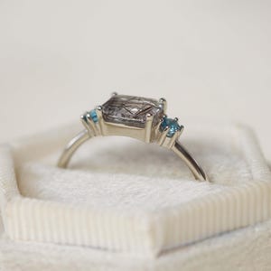 May include: A silver ring featuring a large, square-cut, smoky quartz gemstone with inclusions, flanked by two small, round, light blue gemstones. The ring is set in a delicate, minimalist style and rests in a white velvet ring box.