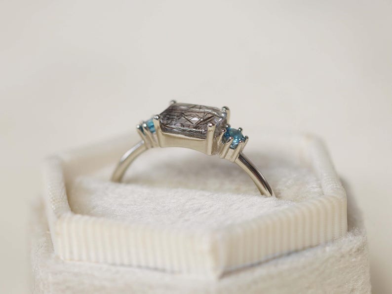 May include: A silver ring featuring a large, square-cut, smoky quartz gemstone with inclusions, flanked by two small, round, light blue gemstones. The ring is set in a delicate, minimalist style and rests in a white velvet ring box.