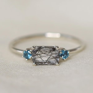 May include: A silver ring featuring a rectangular, clear gemstone with black inclusions, flanked by two smaller, round, blue gemstones. The ring is set against a soft, off-white background, highlighting the jewelry's details.