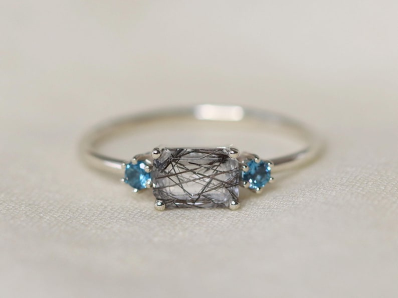 May include: A silver ring featuring a rectangular, clear gemstone with black inclusions, flanked by two smaller, round, blue gemstones. The ring is set against a soft, off-white background, highlighting the jewelry's details.