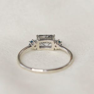 May include: A silver ring with a rectangular, gray-toned gemstone as the centerpiece. Two small, blue gemstones flank each side of the main stone. The ring is set against a white, textured background.