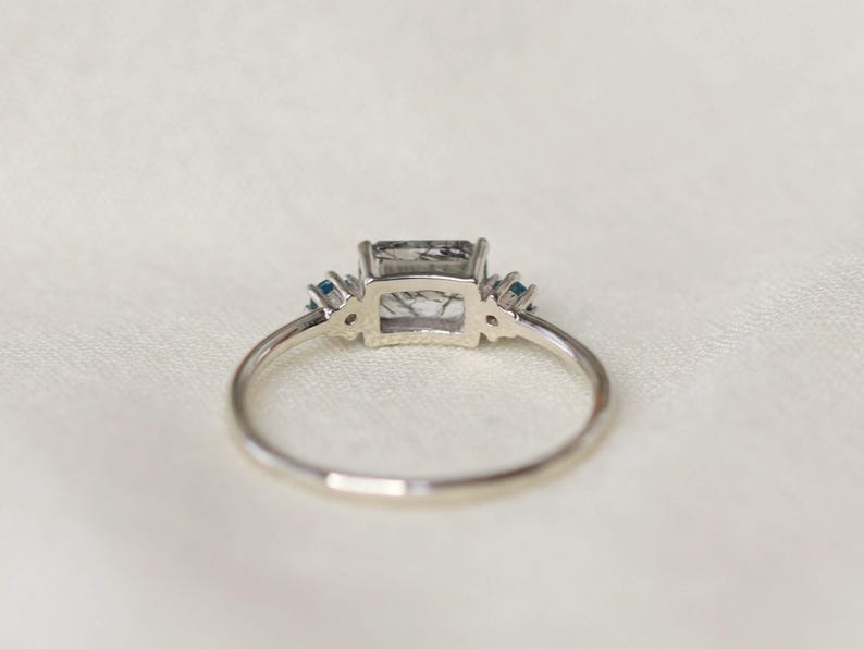 May include: A silver ring with a rectangular, gray-toned gemstone as the centerpiece. Two small, blue gemstones flank each side of the main stone. The ring is set against a white, textured background.