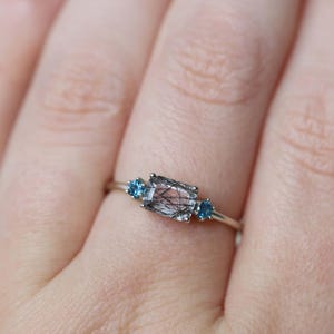 May include: A silver ring featuring a rectangular, clear gemstone with black inclusions, flanked by two small, round, blue gemstones. The ring is set on a delicate silver band, showcasing a minimalist design.