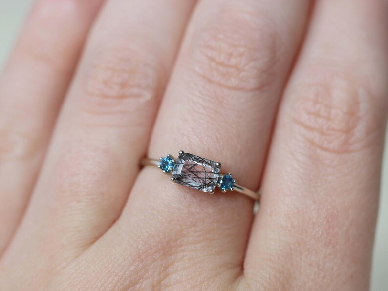 May include: A silver ring featuring a rectangular, clear gemstone with black inclusions, flanked by two small, round, blue gemstones. The ring is set on a delicate silver band, showcasing a minimalist design.