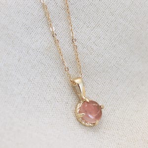 May include: A delicate gold chain necklace with a pink gemstone pendant. The pendant is set in a gold claw setting and has a round shape.