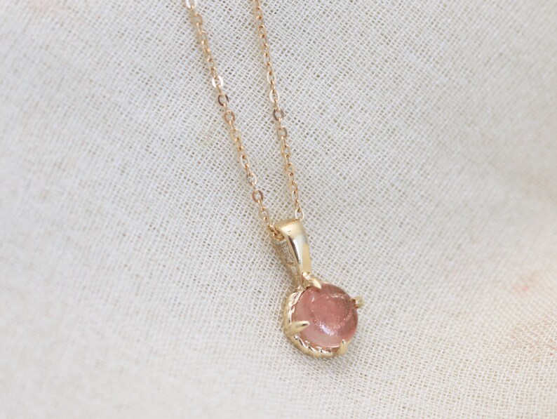 May include: A delicate gold chain necklace with a pink gemstone pendant. The pendant is set in a gold claw setting and has a round shape.