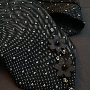 Könnte beinhalten: Eine schwarze Krawatte mit weißem Mikropunktmuster, verziert mit zwei dunkelgrauen blumenförmigen Akzenten und mehreren kleinen, metallisch silberfarbenen blumenförmigen Nieten, die entlang der Stoffkante angeordnet sind.