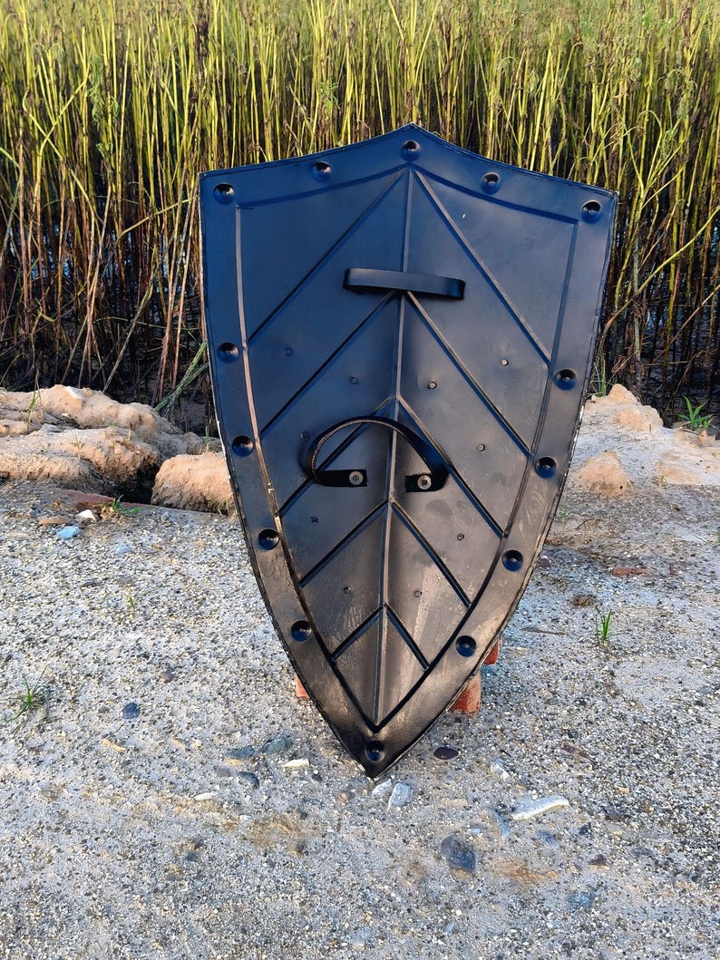 May include: Black plastic shield with a leaf-like design and a handle. The shield has a pointed bottom and is surrounded by rivets. The background includes sand, rocks, and tall grass.