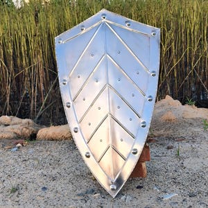 May include: A silver-colored shield with a pointed bottom and rounded top. The shield has a series of diagonal lines and rivets along the edges. The shield is made of metal and is displayed outdoors.