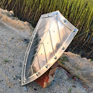 May include: A shiny, silver metal shield with a pointed bottom and a decorative leaf-like pattern. The shield is held up by a red brick, with a background of tall green grass and a sandy ground.