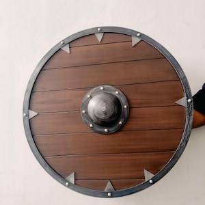 May include: A round wooden shield with a dark brown finish. The shield has a metal rim and a central boss. Triangular metal accents are evenly spaced around the rim. The shield is designed for historical reenactment or display.