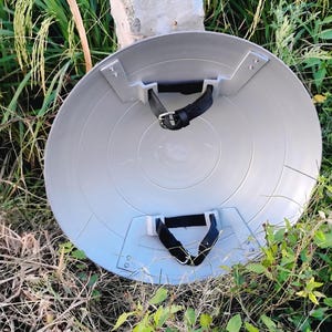 May include: A grey, circular shield with black straps and silver metal brackets. The shield has concentric circles. It is lying on the ground with grass and foliage in the background. The shield is a prop or costume accessory.