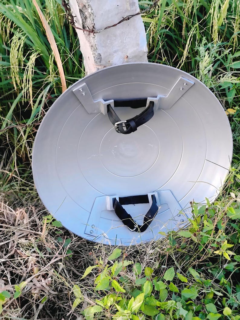 May include: A grey, circular shield with black straps and silver metal brackets. The shield has concentric circles. It is lying on the ground with grass and foliage in the background. The shield is a prop or costume accessory.