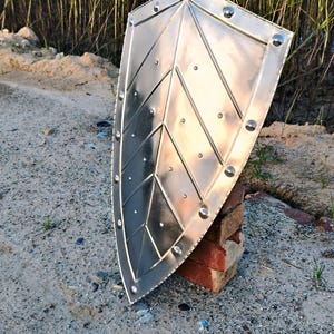 May include: A silver-colored shield with a pointed top and rounded bottom. The shield has a geometric design with rivets along the edges. It is propped up on a stack of red bricks, set against a sandy background.