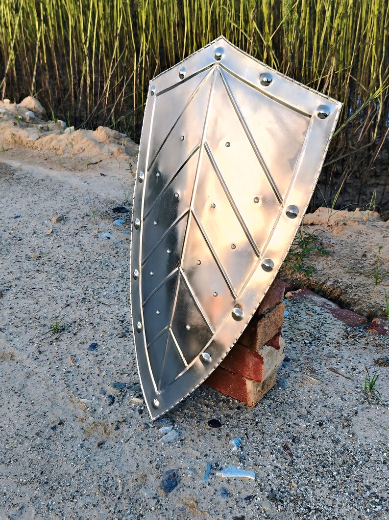 May include: A silver-colored shield with a pointed top and rounded bottom. The shield has a geometric design with rivets along the edges. It is propped up on a stack of red bricks, set against a sandy background.
