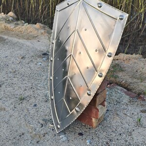 May include: A silver-colored shield with a pointed top and rounded bottom. The shield has a geometric design with rivets along the edges and within the pattern. The shield is propped up on a red brick.