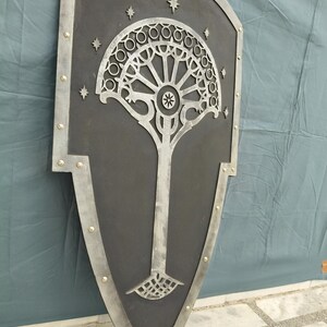 May include: A large, shield-shaped wall hanging with a black background and a silver tree design. The tree has a circular crown and a detailed trunk. The shield is framed with silver and accented with small, gold-colored rivets.