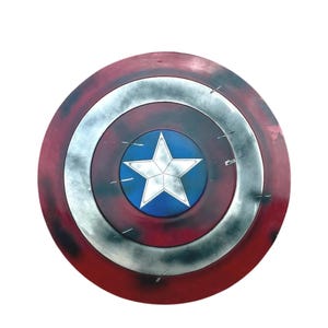 May include: A circular shield featuring a central white star on a blue background, surrounded by a silver ring and an outer red ring. The surface has a weathered, distressed finish with dark smudges and scratches across the red and silver sections.