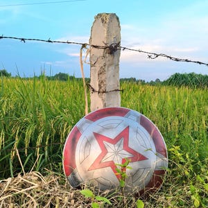 May include: A round shield with a red, white, and silver design featuring a large red star in the center. The shield is resting in tall grass near a concrete post with barbed wire, against a backdrop of a green field and blue sky.
