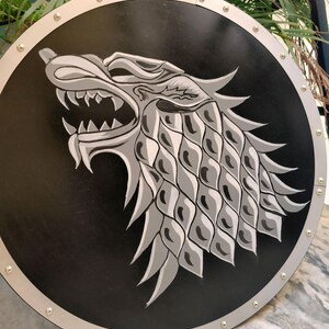 May include: A round, black shield with a silver trim and rivets. The shield features a detailed, gray and white direwolf emblem. The direwolf is snarling, with its head and upper body prominently displayed on the shield's surface.